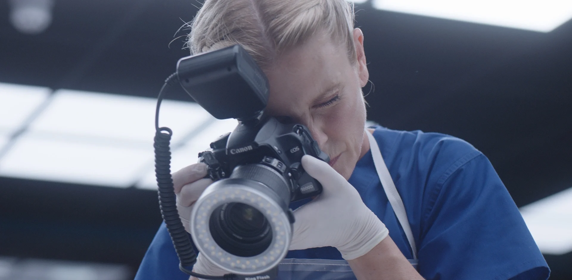 Woman in blue scrubs and gloves photographing with a Canon camera in a well-lit environment.