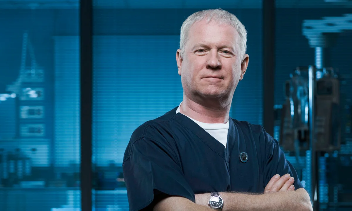 Man in dark scrubs standing with arms crossed, serious expression, in a medical environment with blue tones.