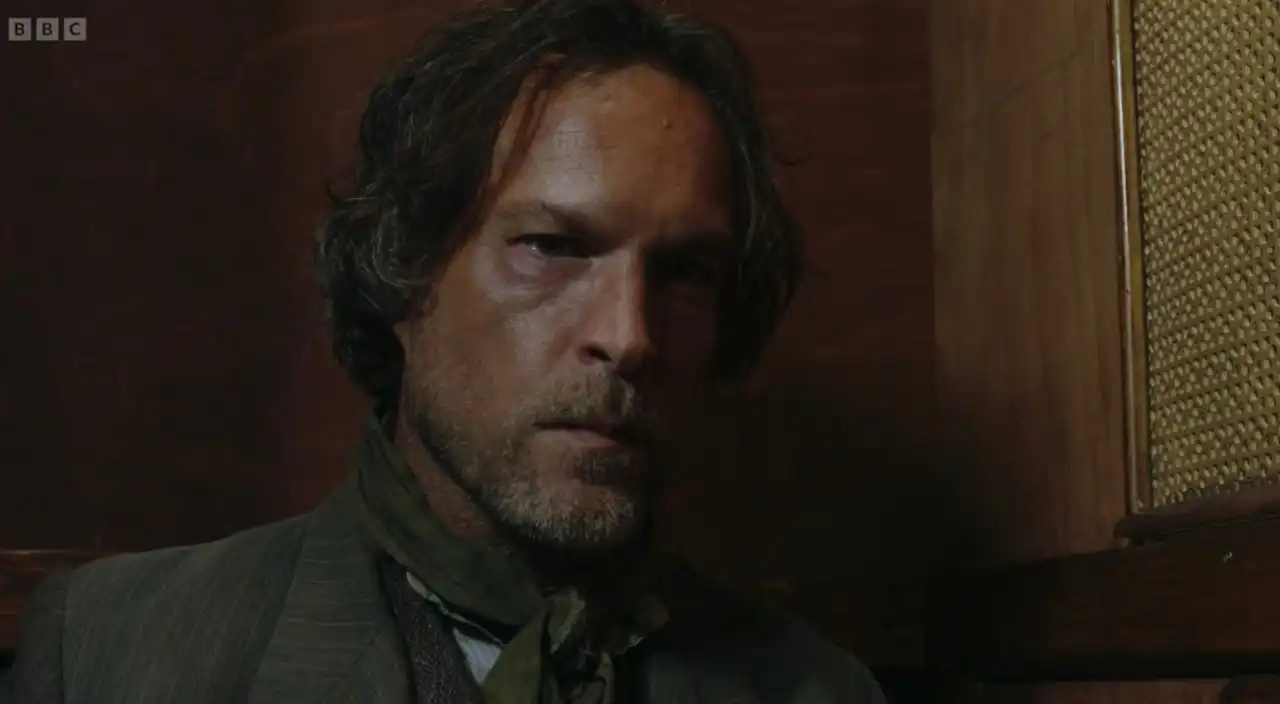 Man with disheveled hair and a scarf, looking serious against a wooden background.