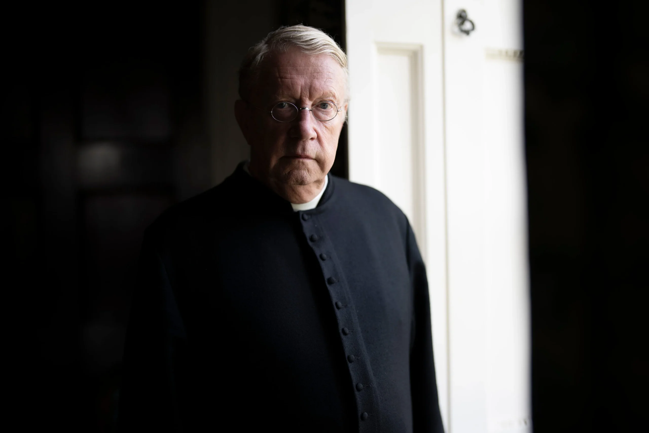 Elderly man in black clergy attire stands by a white door, looking directly at the camera.