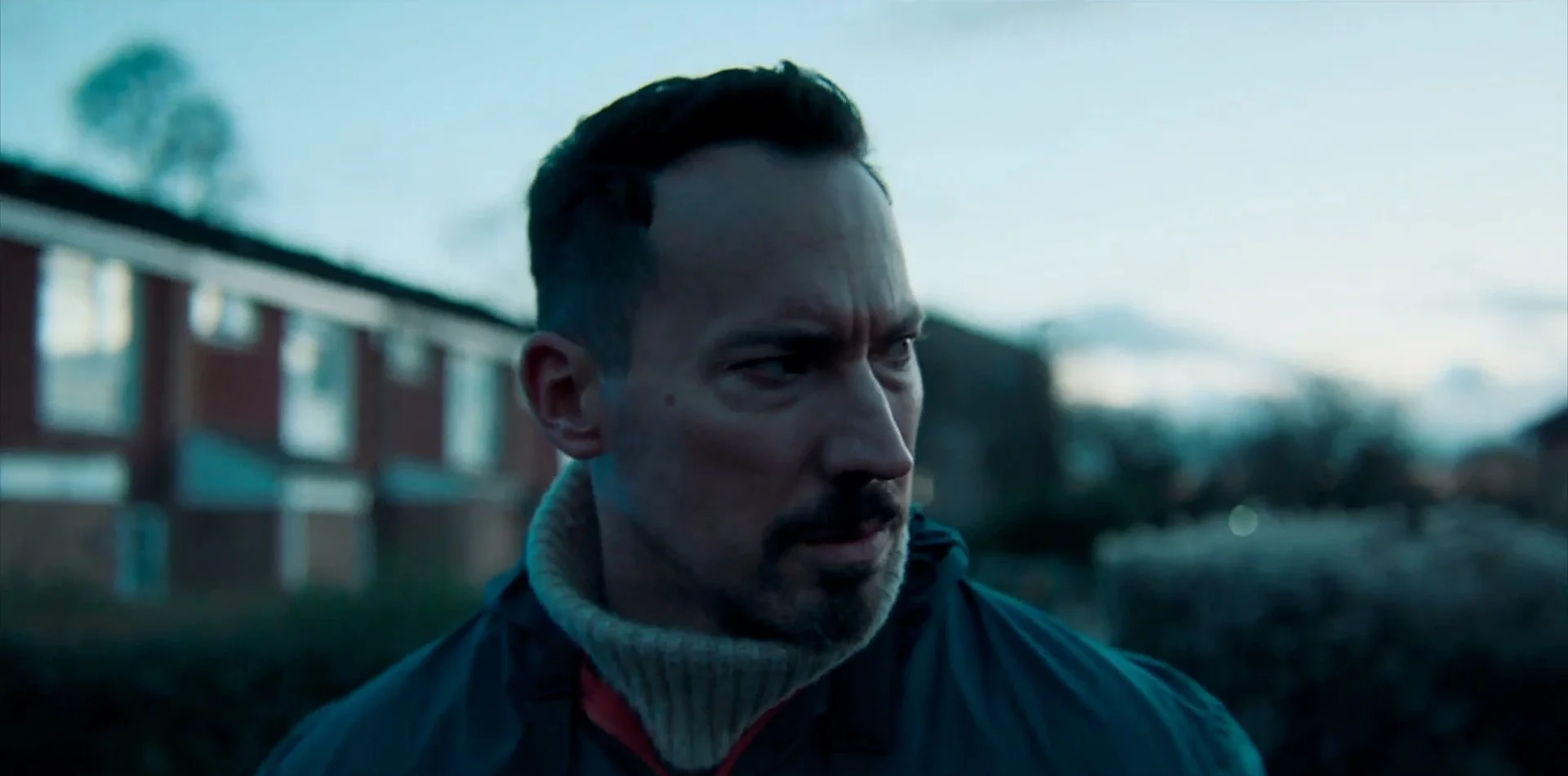 Man with short, dark hair and a sweater looks thoughtfully while standing in a residential area at dusk.